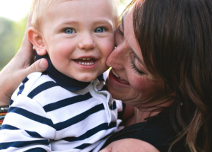 A young child smiles at the camera while being held by his mother who smiles at him while touching her face to his. It is a warm sunny day outside.